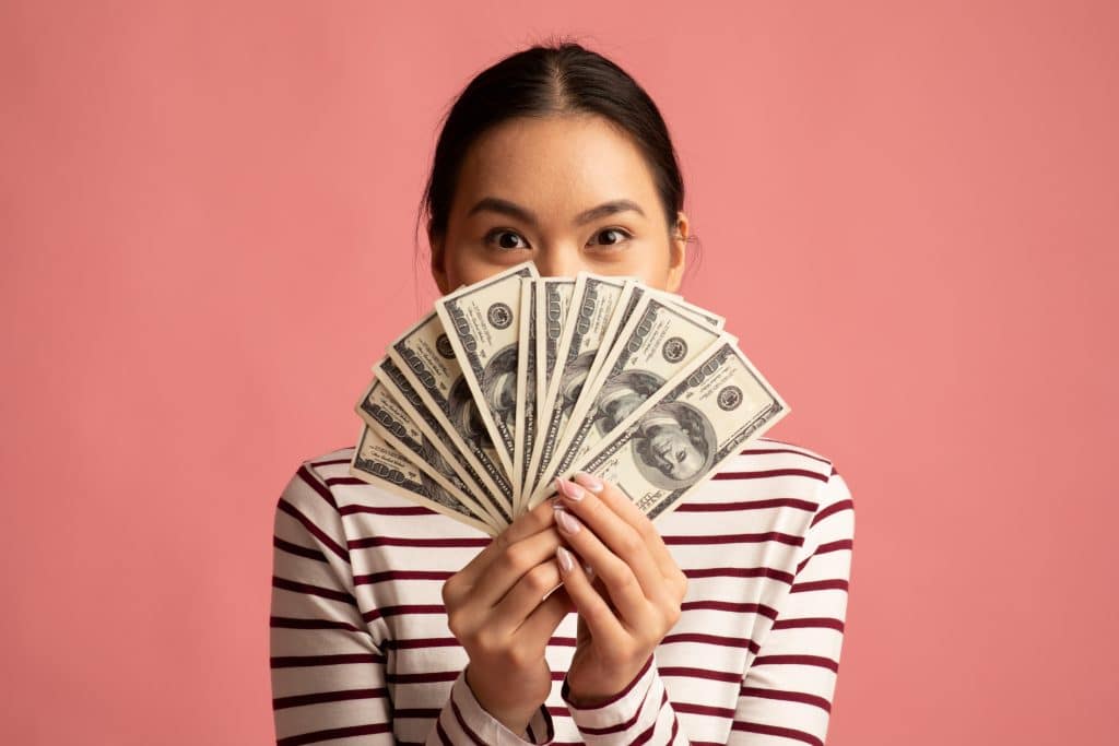 Happy young Asian woman covering face with dollar cash fan
