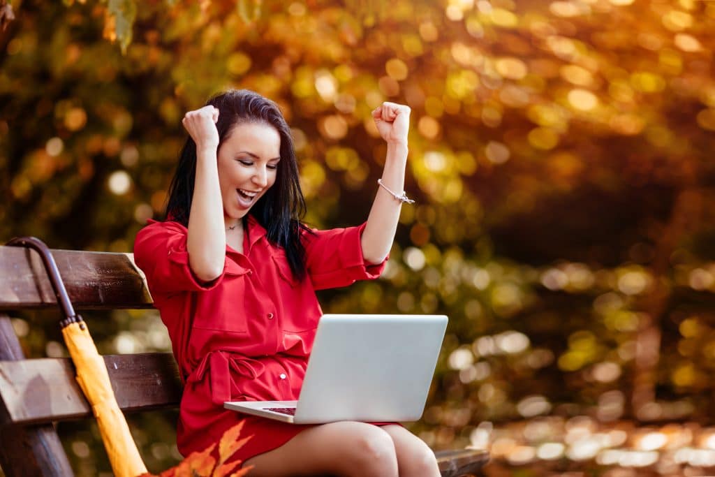Female with laptop sitting on a park bench celebrating job goals